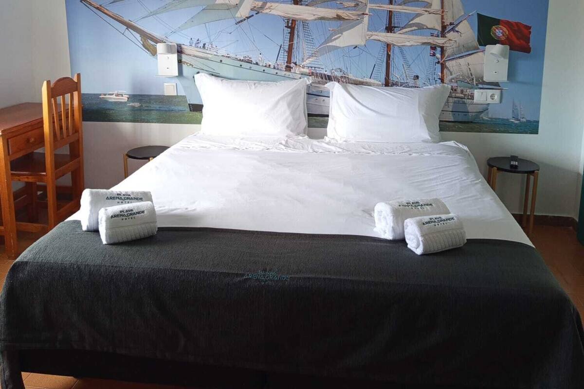 A bed with white linens and towels, a wooden chair, a small table, and a large painting of a sailing ship on the wall.