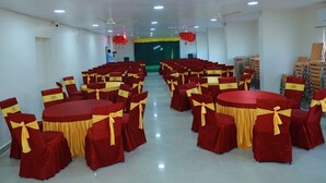 A conference room with round tables and chairs, ceiling fans, and a stage with red decorations. - samudra inn (Nalgonda)