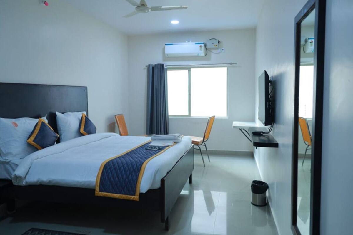 A hotel room with a bed, a desk, a chair, an air conditioning unit, and a television.