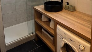 A modern bathroom with a glass-enclosed shower, a round mirror with LED lights, a black sink basin, a soap dispenser, and a washing machine.