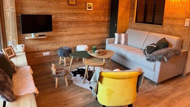 A cozy living room with a wooden wall, a grey sofa, a yellow chair, a small round table, and a TV mounted on the wall.