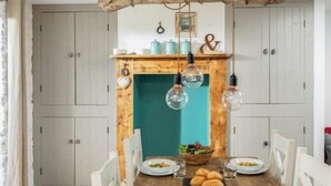 A dining area with a wooden table set for four, surrounded by white chairs. There are hanging light bulbs above the table, and a teal-colored wall behind it.