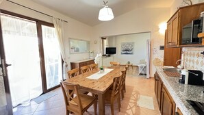 A well-lit kitchen with wooden cabinets, a dining table, and chairs. There is a TV on the wall and a painting above it. - Terraced house with swimming pool, surrounded by nature, 250 meters from the sea (Marina, Tertenia)