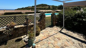 A stone patio with a pergola structure, a pool in the background, and a view of terraced houses. - Terraced house with swimming pool, surrounded by nature, 250 meters from the sea (Marina, Tertenia)