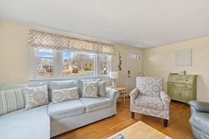 A living room with a sofa, armchair, wooden floor, window with view, door, and a cabinet. - Sandcastles & Sunsets: A Cape Cod Family Haven (Yarmouth)