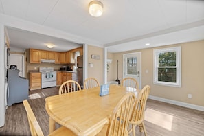 A dining area with a wooden table and chairs, a kitchen in the background, and a window with a view.