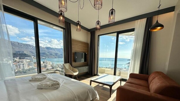 Quarto de hotel com cama, sofá, televisão e amplas janelas com vista para a cidade e para o mar.