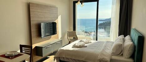 A hotel room with a bed, a desk, a chair, a TV, and a large window with a view of the sea.