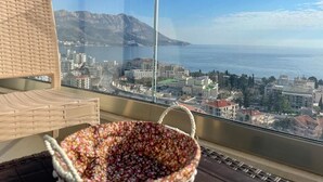 Varanda com cadeira de vime e cesto de flores, com vista para uma paisagem costeira e montanhas ao fundo.