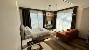 Quarto de hotel moderno com cama, área de estar com sofá e poltrona, mesa de jantar com cadeira e televisão na parede.