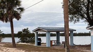 A covered pavilion with a picnic table and trash bin, located near a body of water with trees and a sandy beach.
