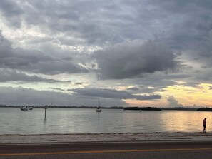 A waterfront view with boats, a person standing by the shore, and a road in the foreground.