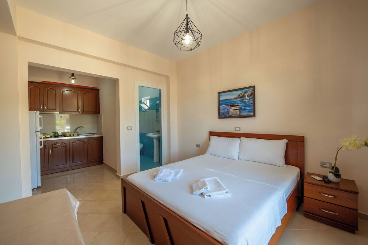 A bedroom with a large bed, a nightstand, a painting, and a view of the bathroom and kitchen.