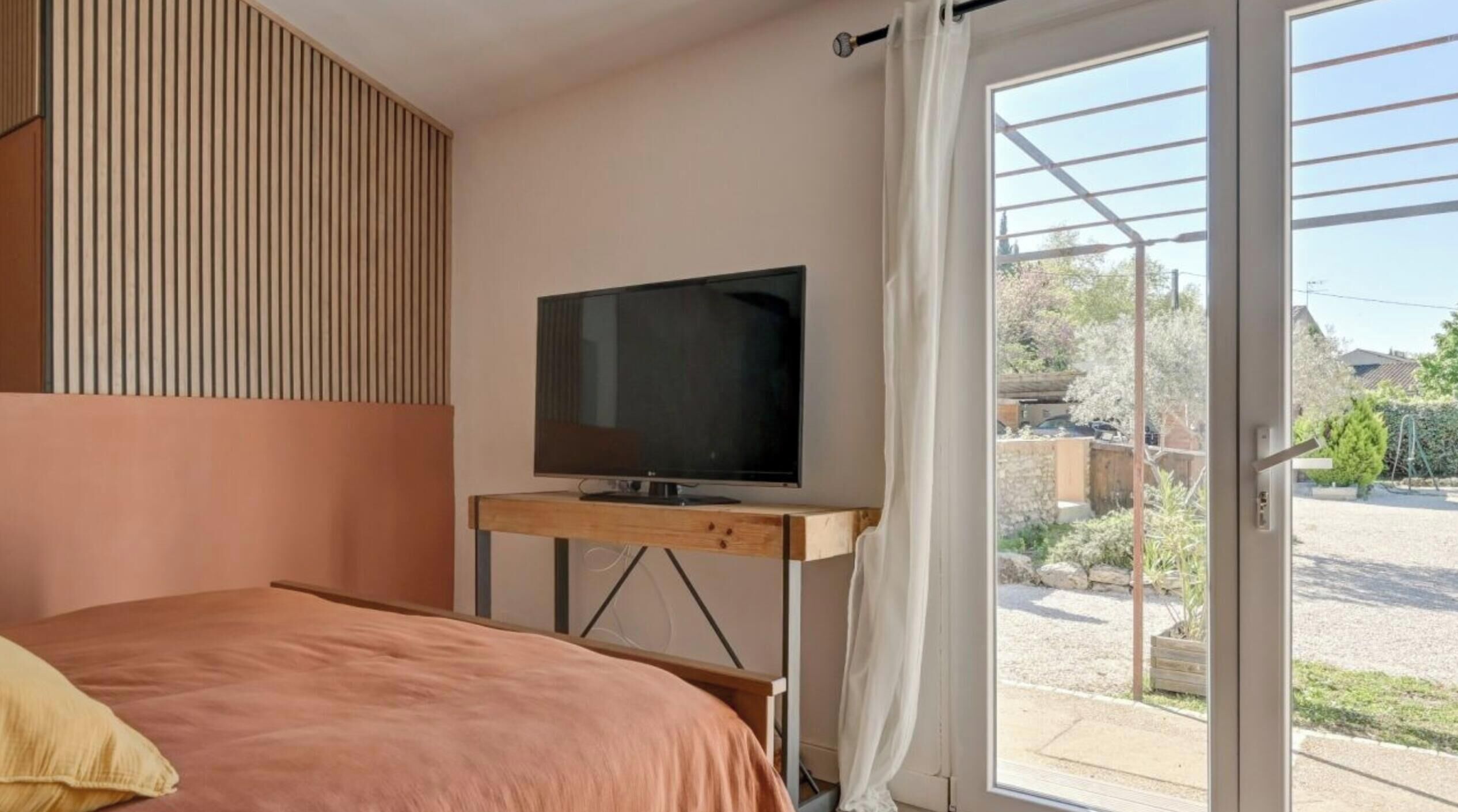 A bedroom with a bed, a TV on a wooden stand, and a sliding glass door leading to an outdoor area.