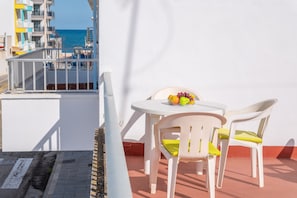 Un balcon avec une table et des chaises blanches, une assiette de fruits, et une vue sur la mer et les bâtiments en arrière-plan.