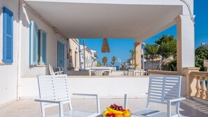 A terrace with white chairs and a table, a bowl of fruit, and a view of the street and buildings. - Can Calpe in Mallorca (Santa Margalida)