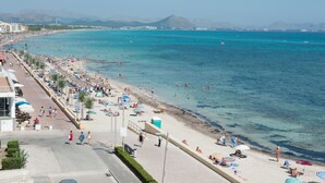 Una platja amb aigua blava cristal·lina, un passeig marítim amb ombrel·les i edificis al fons.