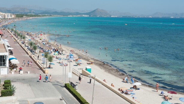 A beach with clear blue water, a promenade with umbrellas, and buildings in the background. - Can Calpe in Mallorca (Santa Margalida)