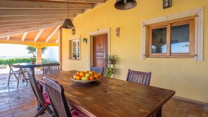 A dining area with a wooden table, chairs, and a bowl of fruit. There are windows and a door in the background. - Casa Judith in Mallorca (Muro)