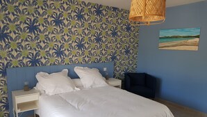 A bedroom with a bed, white pillows, a nightstand, a lamp, a chair, and a tropical wallpaper. - Les Terrasses d'Arutoli (Porto-Vecchio)