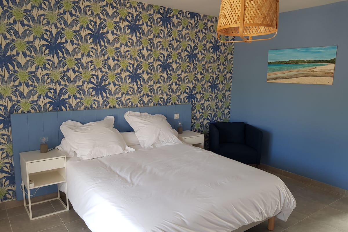 Een slaapkamer met een bed, witte kussens, een nachtkastje, een lamp, een stoel en tropische behang.