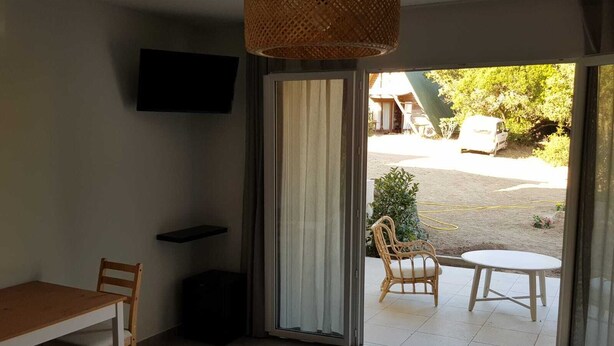 A room with a bed, a table, and chairs, a TV on the wall, and a sliding glass door leading to an outdoor area with a table and chairs. - Les Terrasses d'Arutoli (Porto-Vecchio)