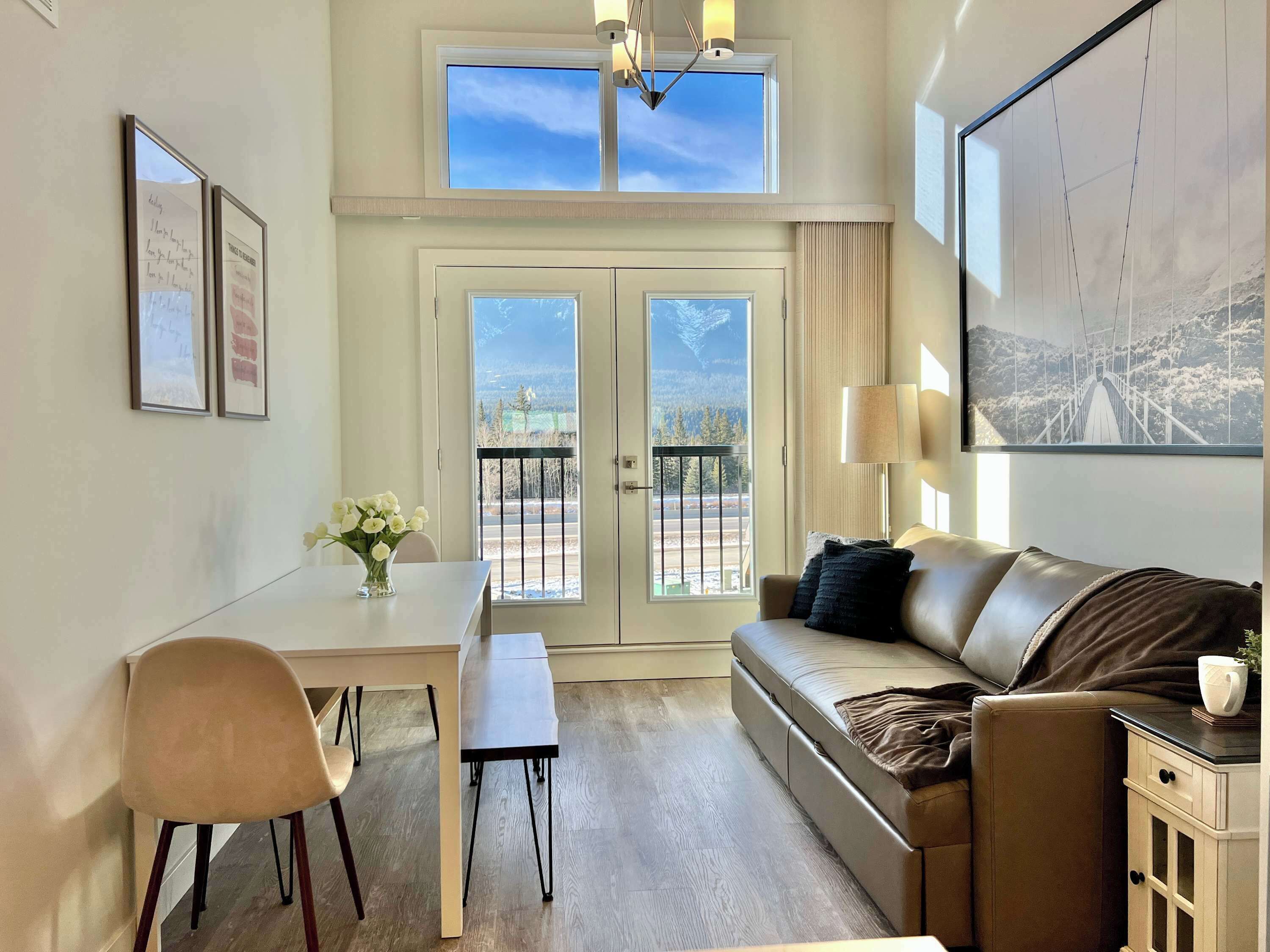 Una habitación con una mesa de comedor, un sofá, una silla, un jarrón de flores y vistas a montañas desde la puerta.