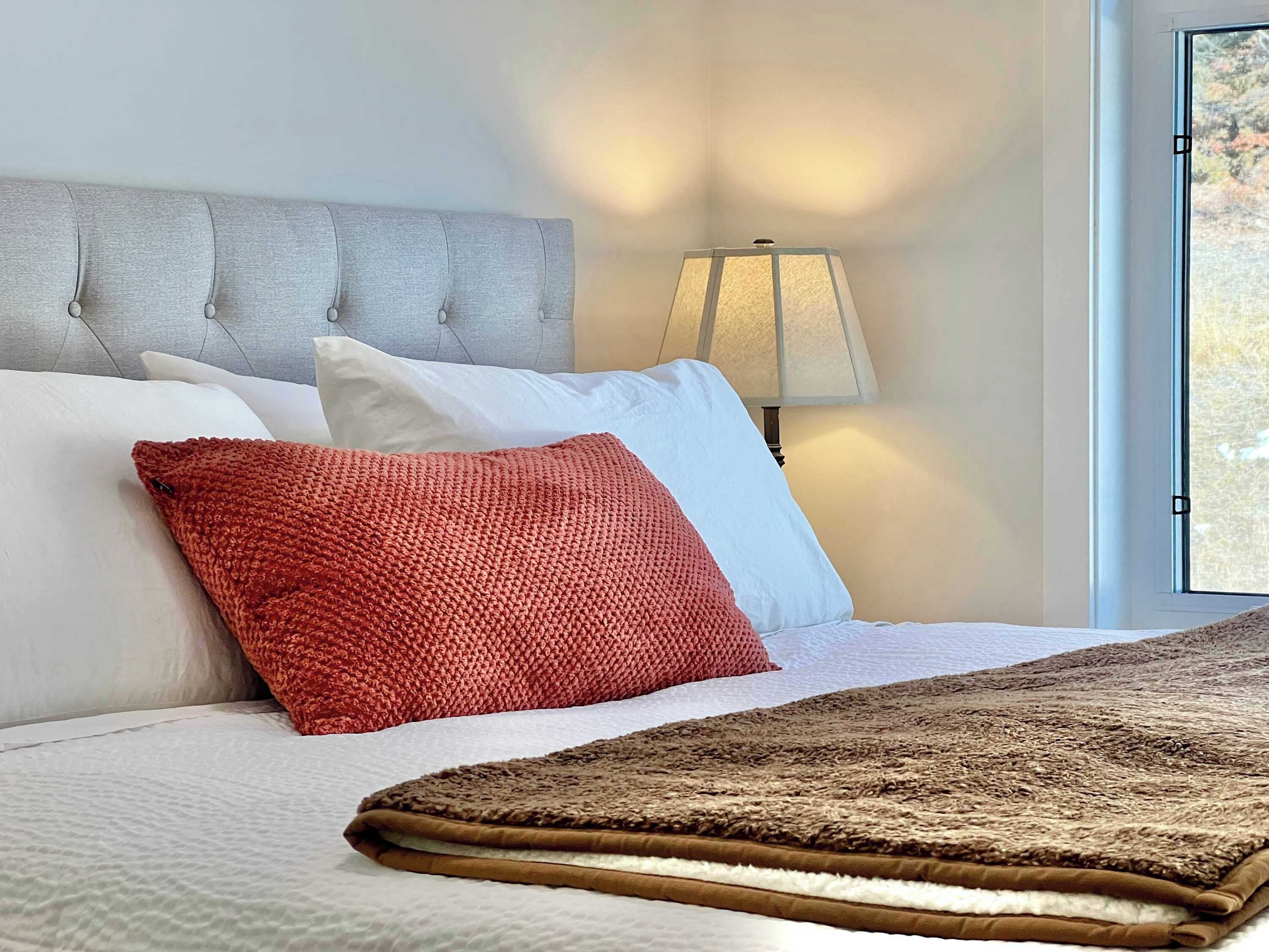 A neatly made bed with a red pillow, white pillows, and a brown blanket.