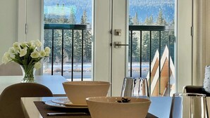 A room with a dining table, chairs, a vase of flowers, and a view of snow-capped mountains through large windows.