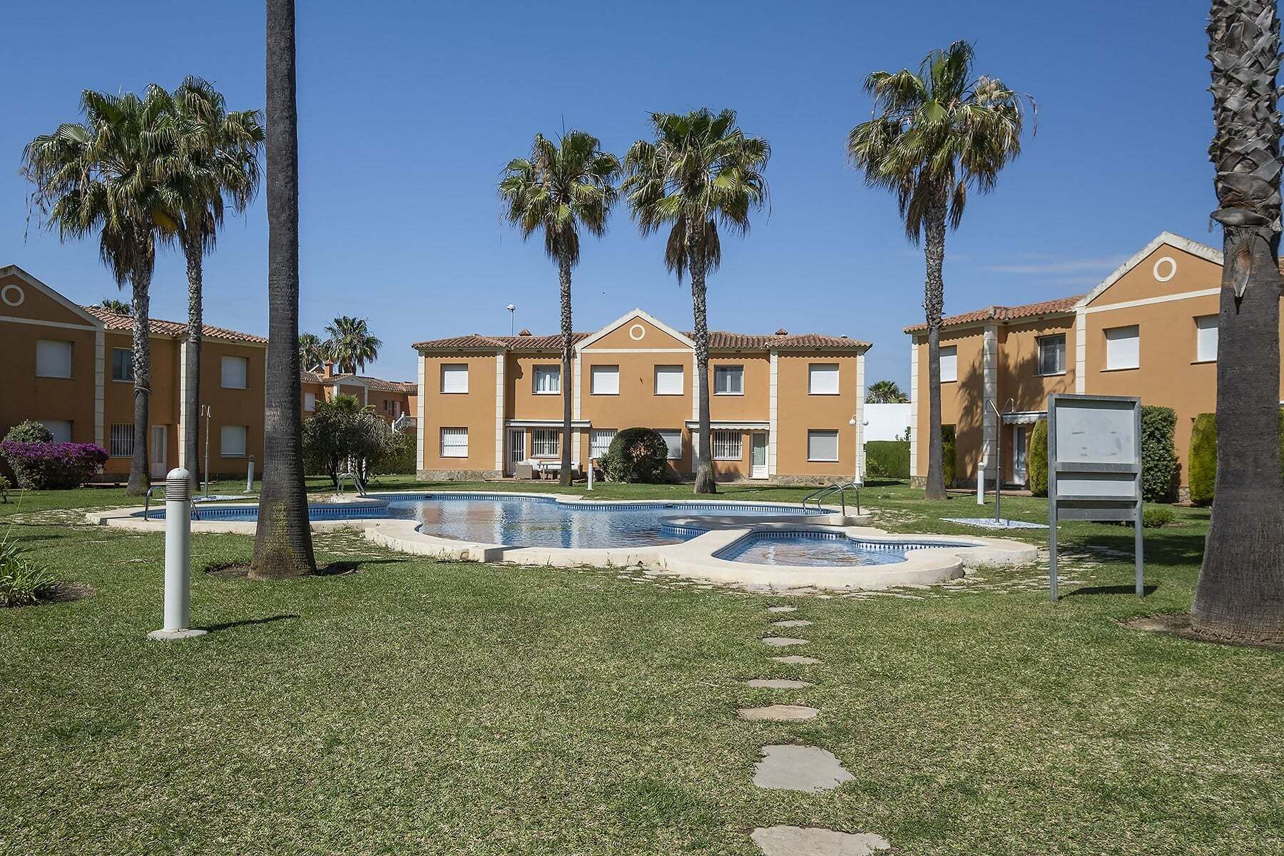 A pool area with palm trees, a clear sky, and a building with a tiled roof.