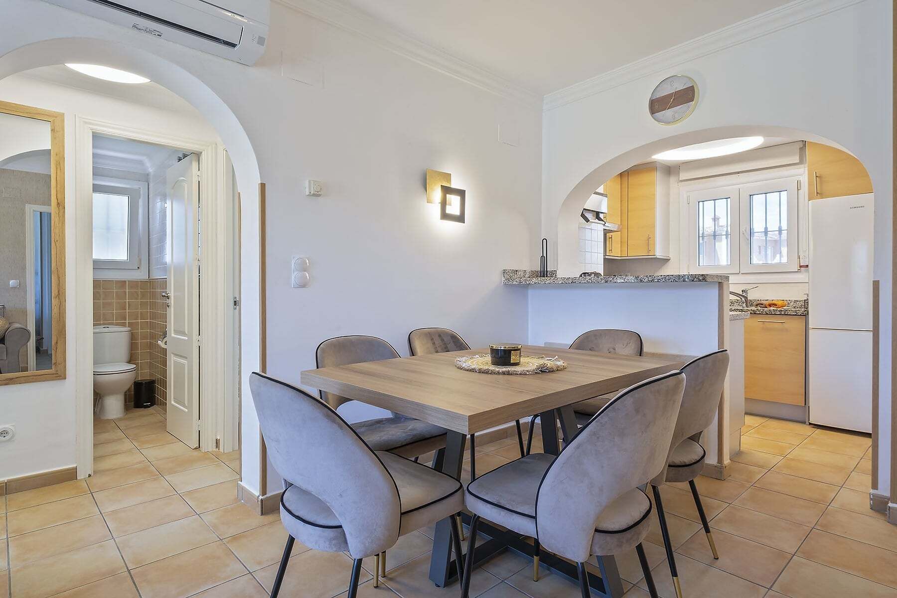 A dining area with a wooden table and six chairs, an air conditioning unit, a kitchen with a refrigerator, and a clock on the wall.