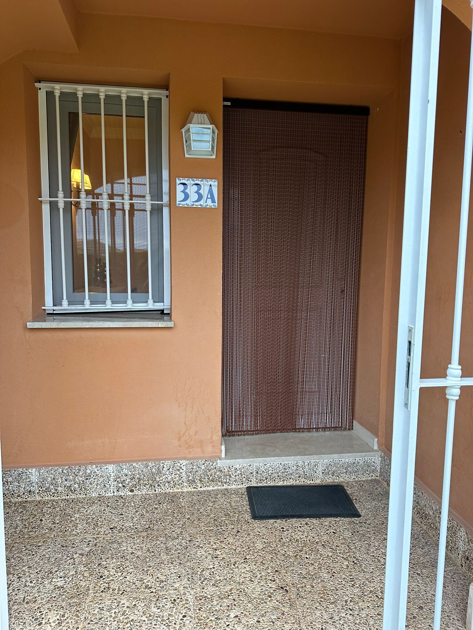 Ein Gebäudeeingang mit vergittertem Fenster, einer Tür mit strukturierter Oberfläche und einem Schild mit der Aufschrift '33A' an der Wand.