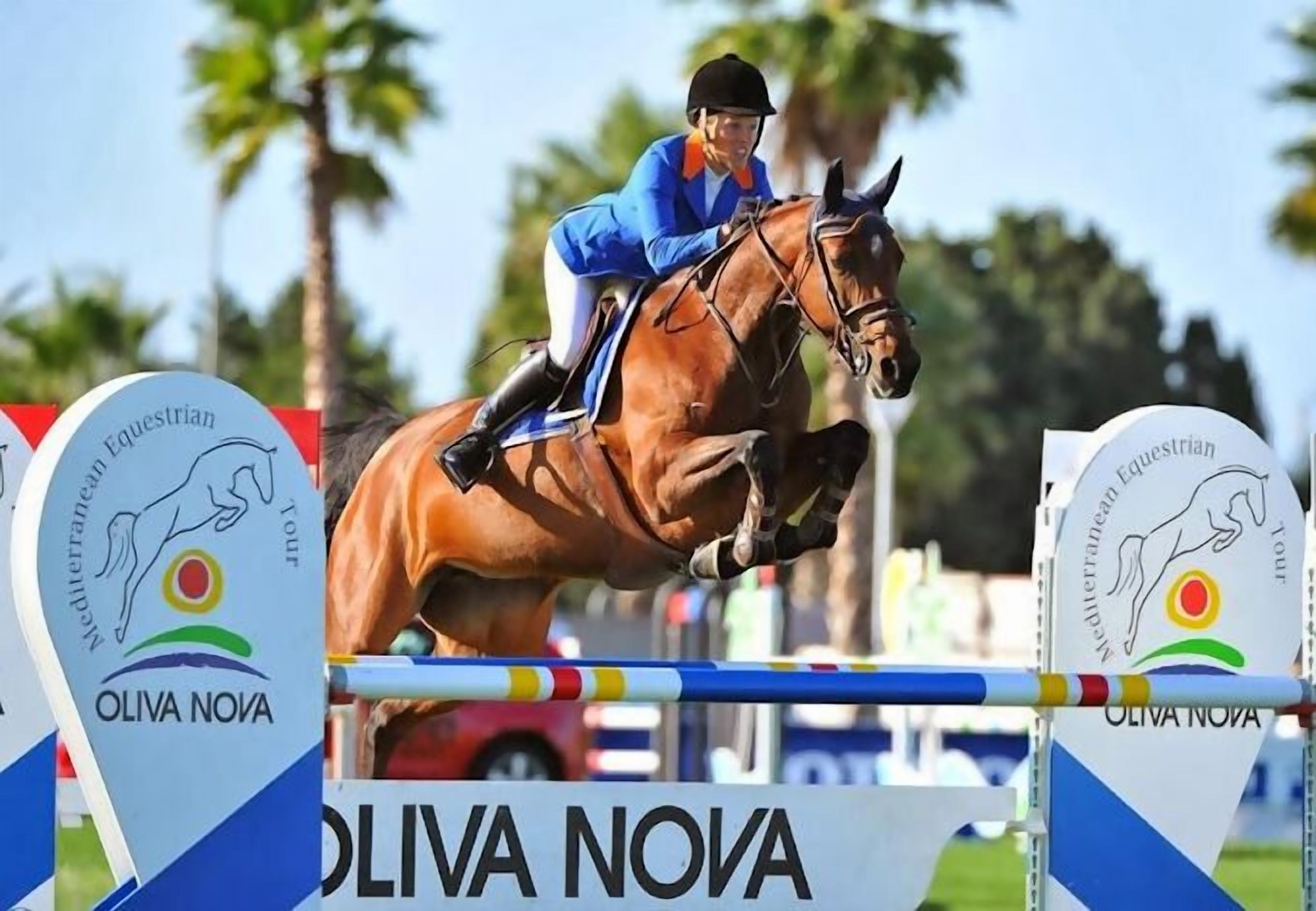 A horse and rider jumping over an obstacle with signs labeled "Oliva Nova" and "Mediterranean Equestrian Tour".