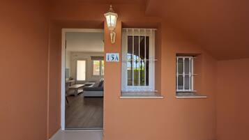 A hallway with a door leading to a living room, a window with a number sign, and a wall-mounted light fixture.