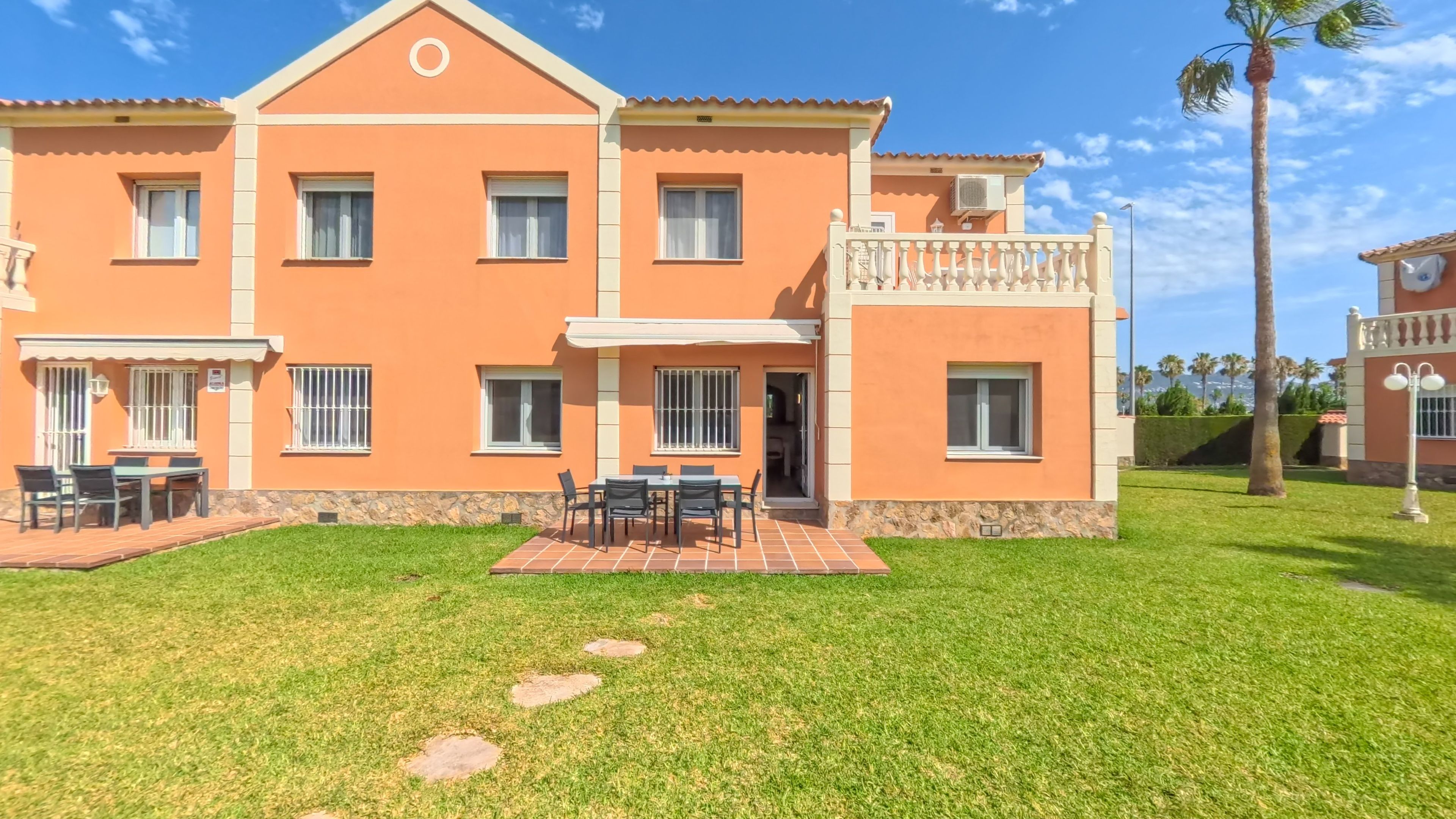Edificio de dos pisos con balcón, patio con mobiliario exterior y césped cuidado.