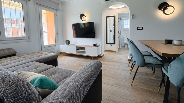 Una sala de estar moderna con televisor de pantalla plana, comedor con mesa y sillas de madera, y vistas al exterior a través de amplios ventanales.