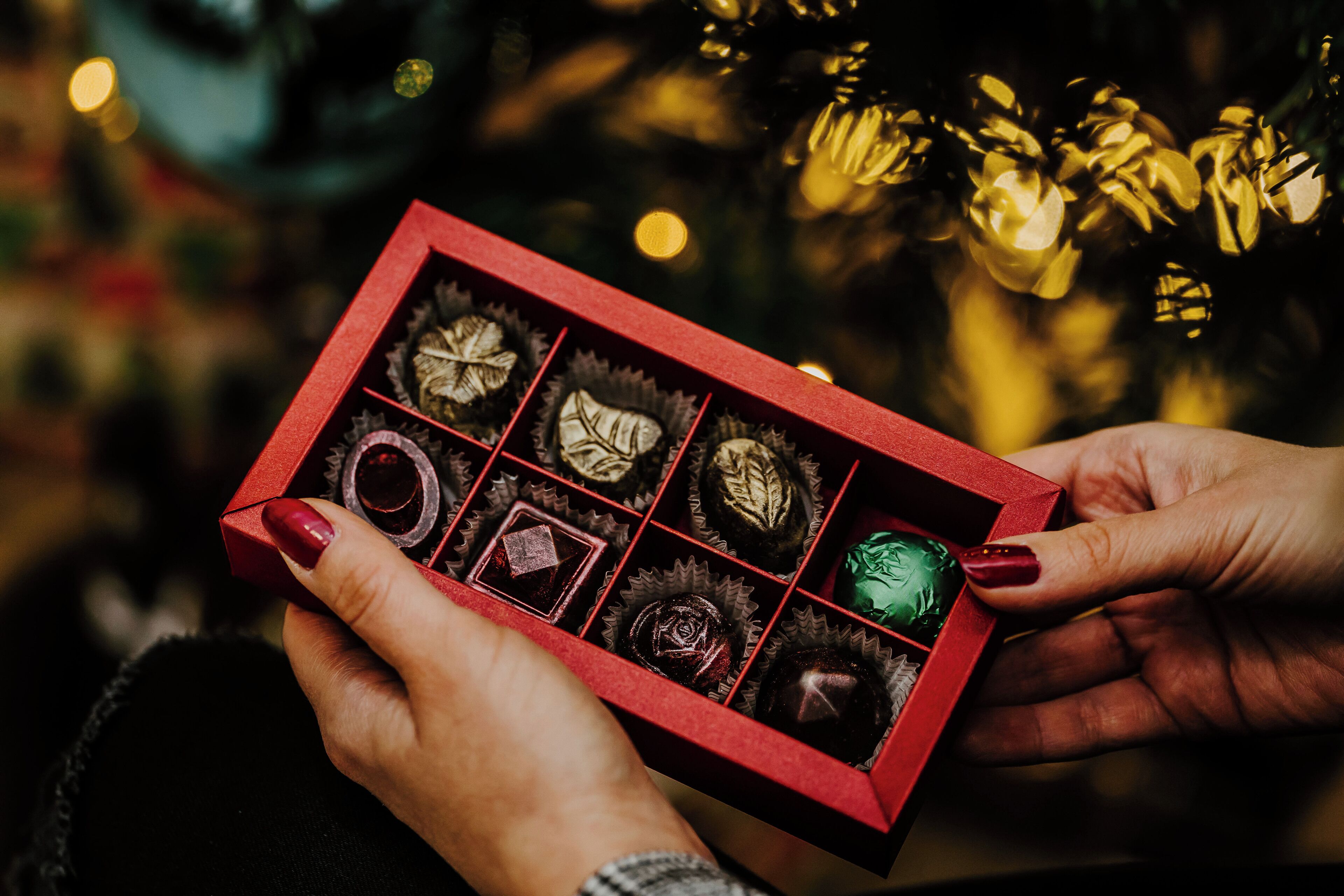A person holding a red box of chocolates with a blurred Christmas tree in the background.