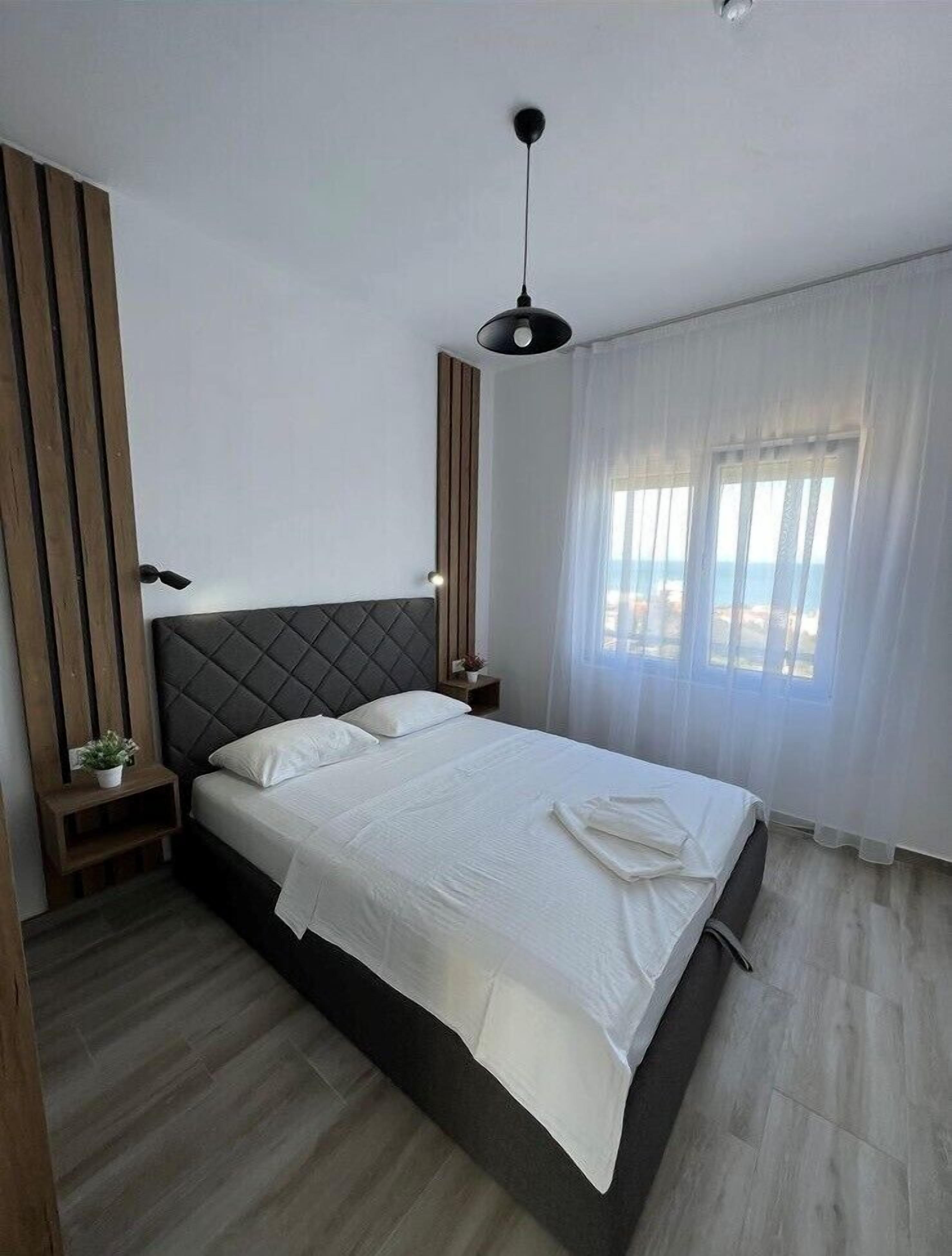 A modern bedroom with a large bed, wooden paneling on the wall, a ceiling light, and a view of the sea through sheer curtains.