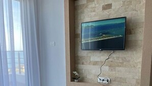 A modern living room with a flat-screen TV mounted on a stone wall, a round wooden coffee table with remote controls, and a view of the sea through sheer curtains. - NEW! Apartment 3 with Pool in Bar, Montenegro (Bar)