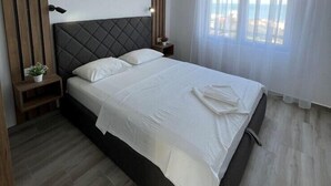 A modern bedroom with a large bed, wooden paneling on the wall, a ceiling light, and a view of the sea through sheer curtains. - NEW! Apartment 4 with Pool in Bar, Montenegro (Bar)