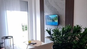 A modern living room with a wooden floor, a potted plant, a flat-screen TV mounted on the wall, and a large window with sheer curtains. - NEW! Apartment 4 with Pool in Bar, Montenegro (Bar)