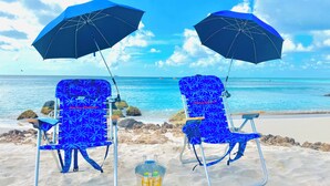 Two beach chairs with blue umbrellas and a bucket of drinks on the sand.