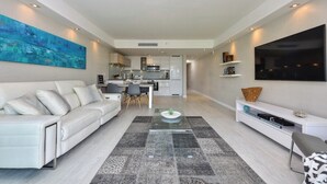 A modern living room with a sofa, coffee table, and TV. There is a kitchen in the background with a dining area.