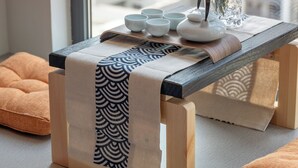 Uma mesa de madeira com um bule branco e chávenas, um vaso com flores e um tecido estampado.