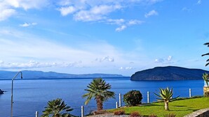 A coastal landscape with a clear view of the sea, a distant mountain, and a well-maintained grassy area with palm trees. - Luxurious Villa Tholos Crete (Ierapetra)