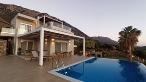 A modern two-story house with a pool, outdoor seating, and a mountain backdrop. - Luxurious Villa Tholos Crete (Ierapetra)