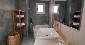 A modern bathroom with a large bathtub, a white sink, and a mirror above it. There are wooden shelves with towels on the left and a wicker basket on the floor. - Luxurious Villa Tholos Crete (Ierapetra)