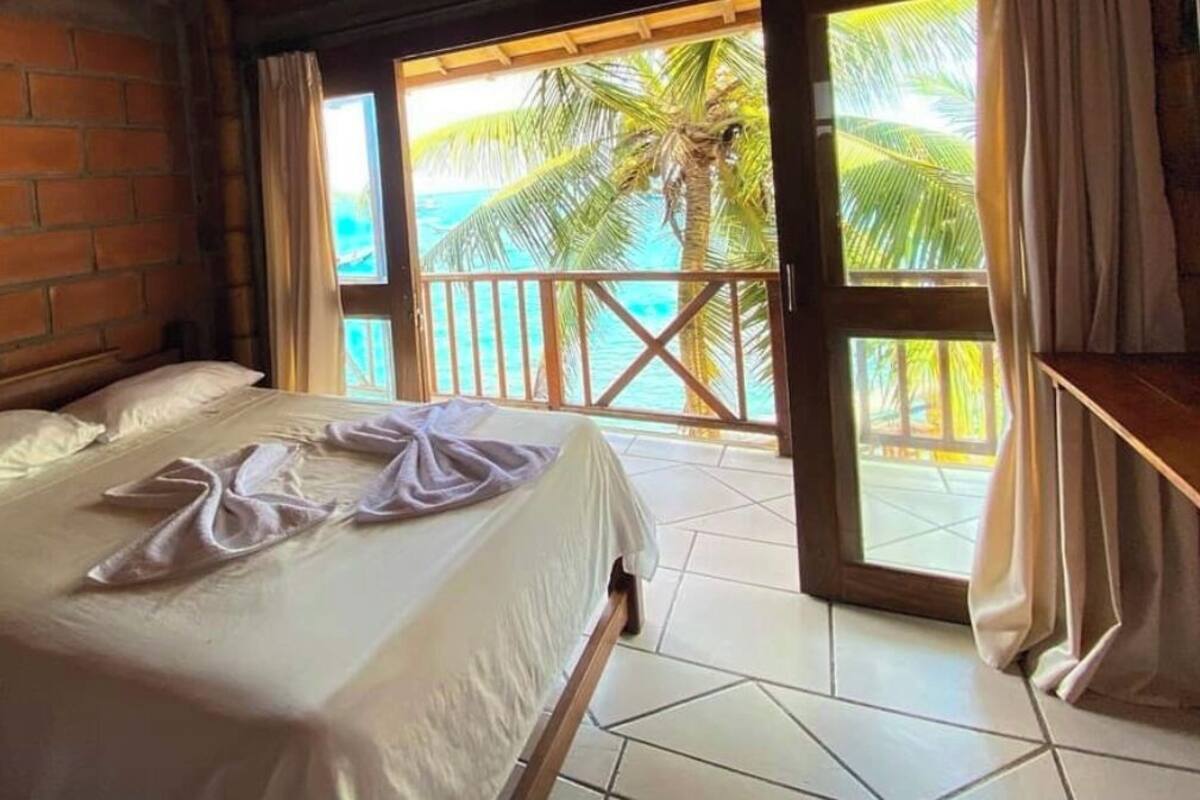 A bedroom with a bed, a wooden headboard, a balcony with a view of the sea and palm trees.