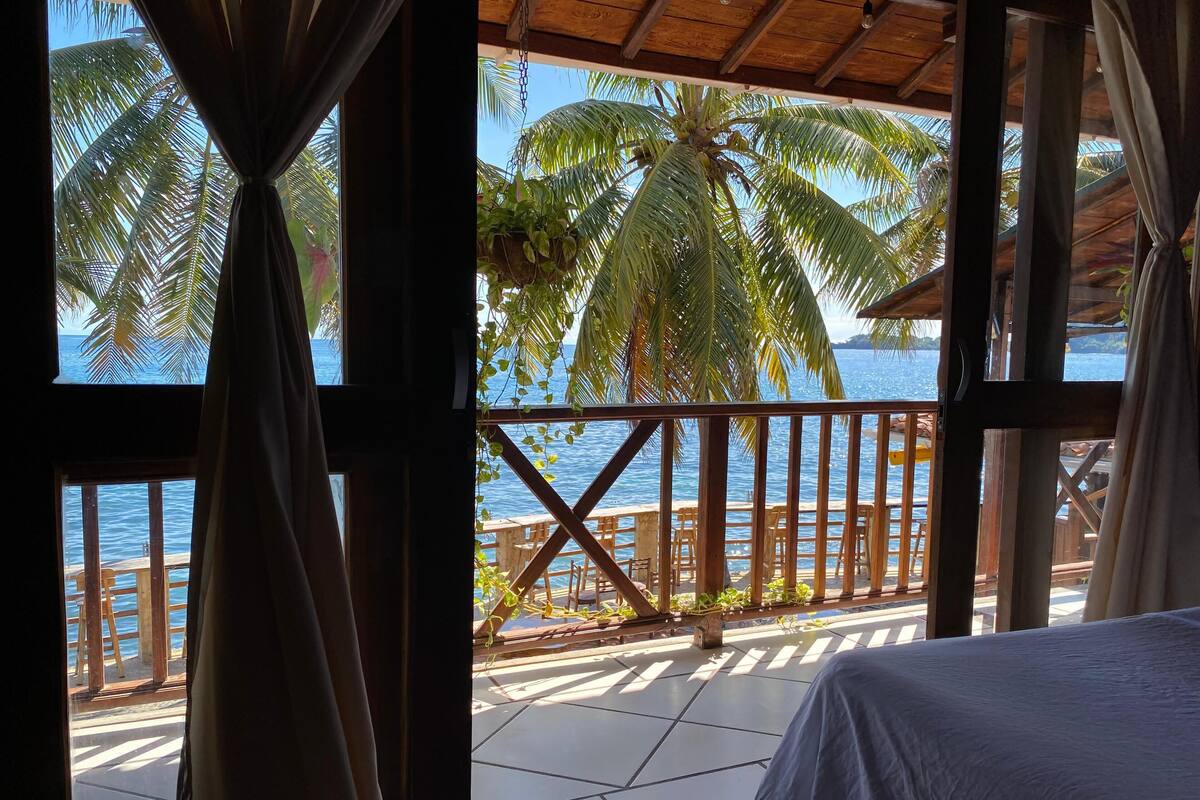 A balcony with a wooden railing, a bed, and a view of the sea and palm trees.
