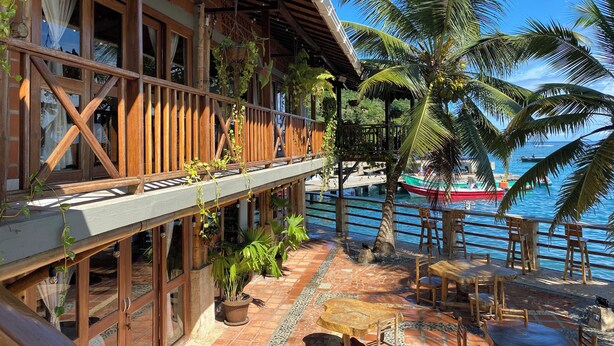 A wooden building with a balcony overlooking a beach, featuring potted plants and outdoor seating.